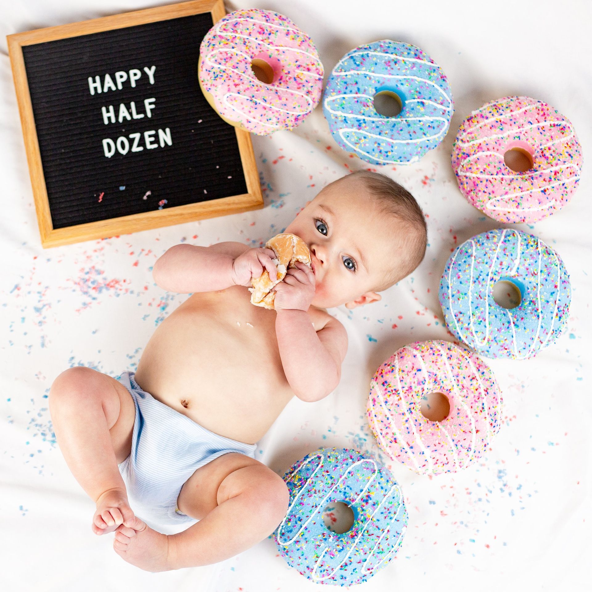 A baby is laying on a bed with donuts and a sign that says `` happy half dozen ''.