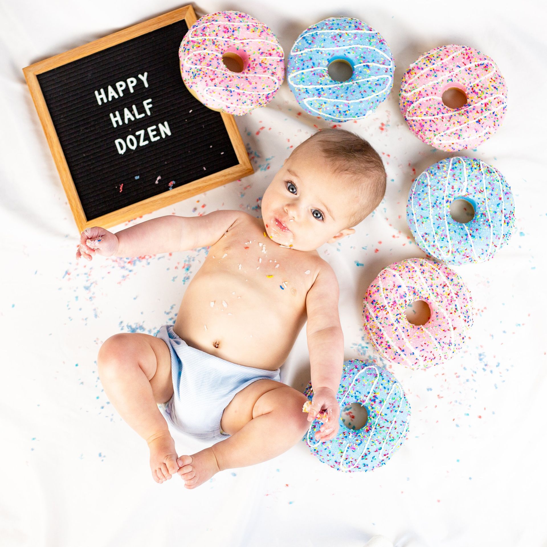 A baby is laying on a bed surrounded by donuts and sprinkles.