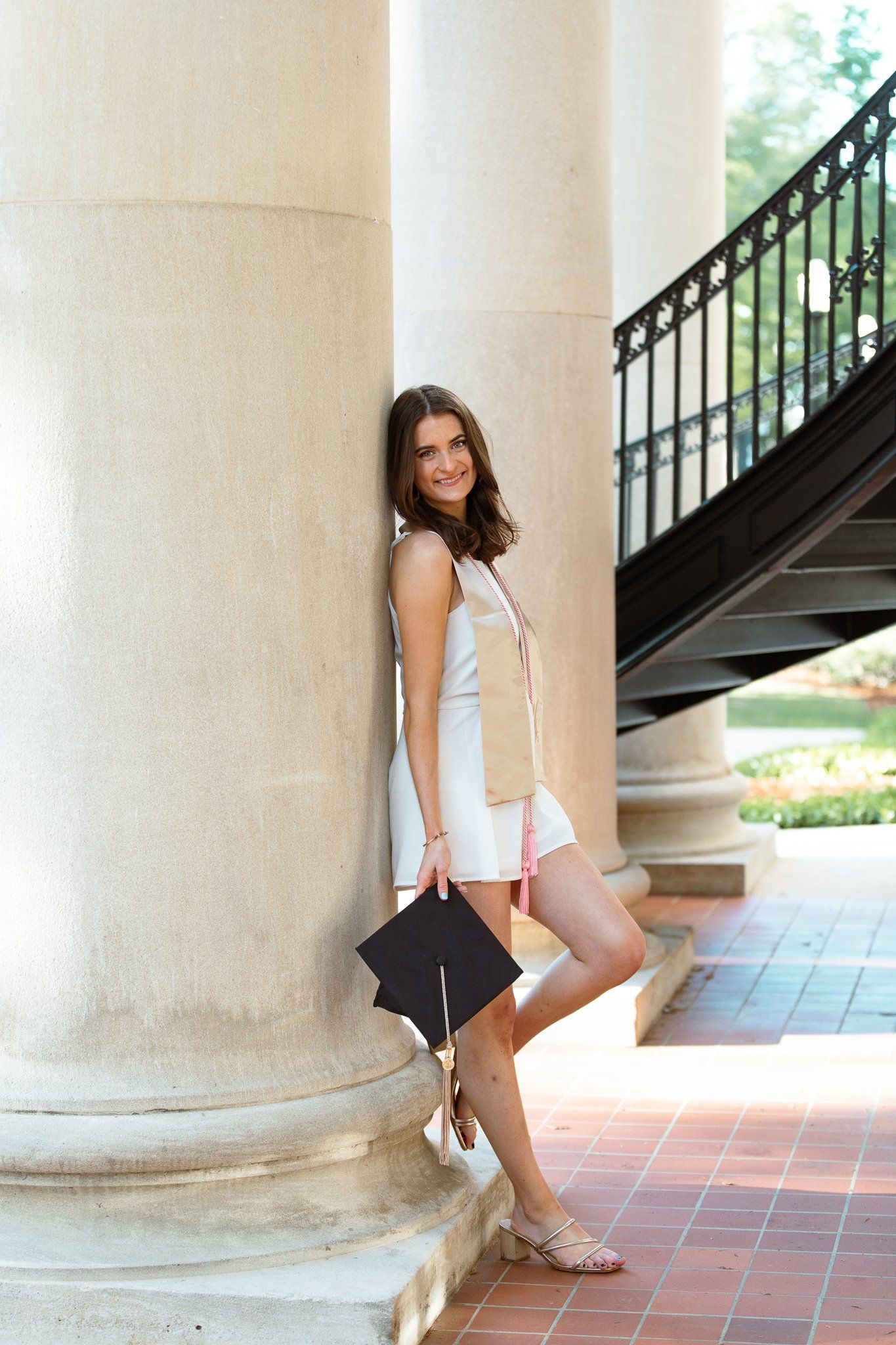 A woman leaning against a pillar holding a graduation cap