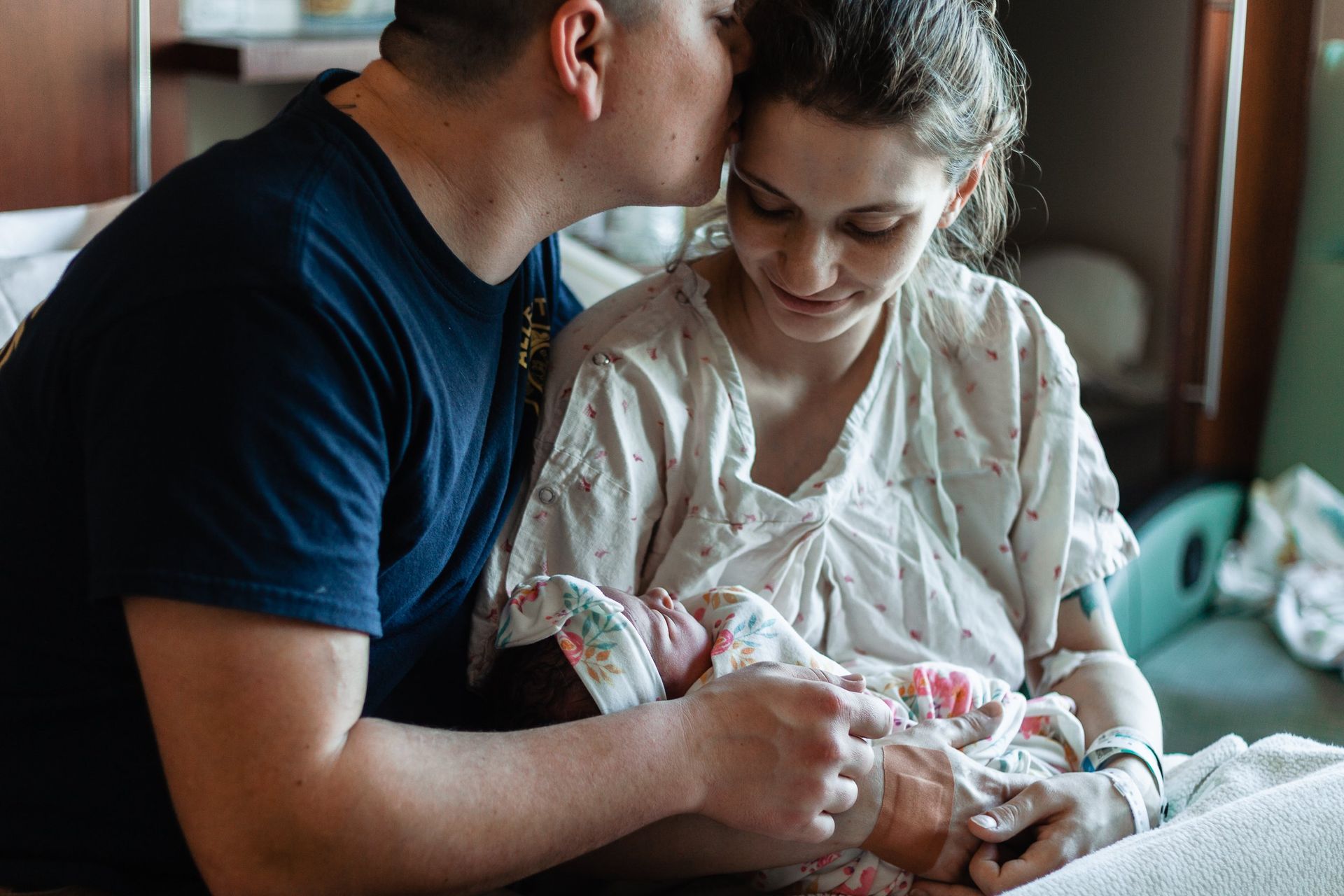 A man is kissing a woman on the cheek while holding a newborn baby in a hospital room.