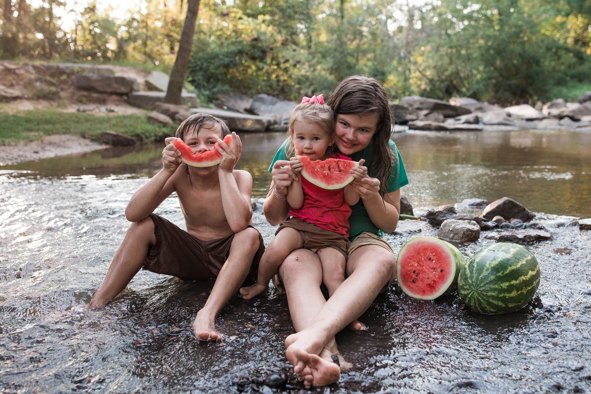 A woman and two children are eating watermelon by the river.