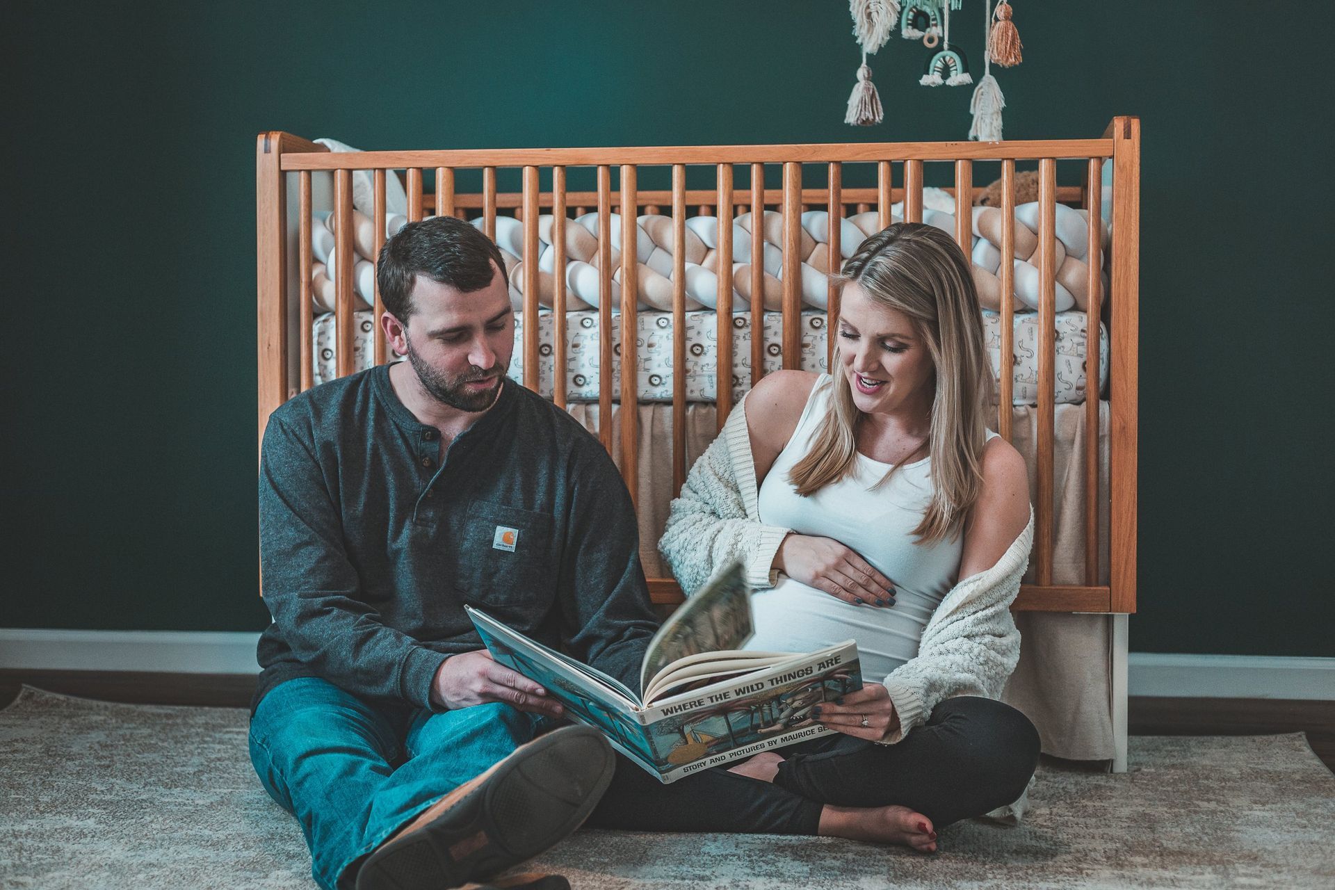 A man and a pregnant woman are sitting on the floor reading a book in front of a crib.