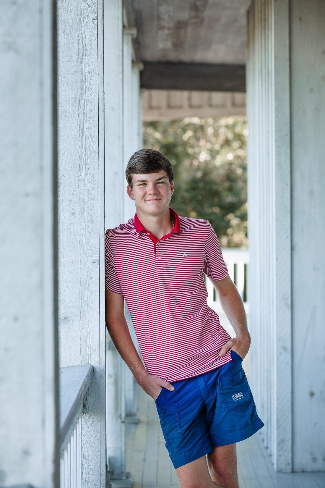 A young man in a red and white striped polo shirt and blue shorts is leaning against a white wall.