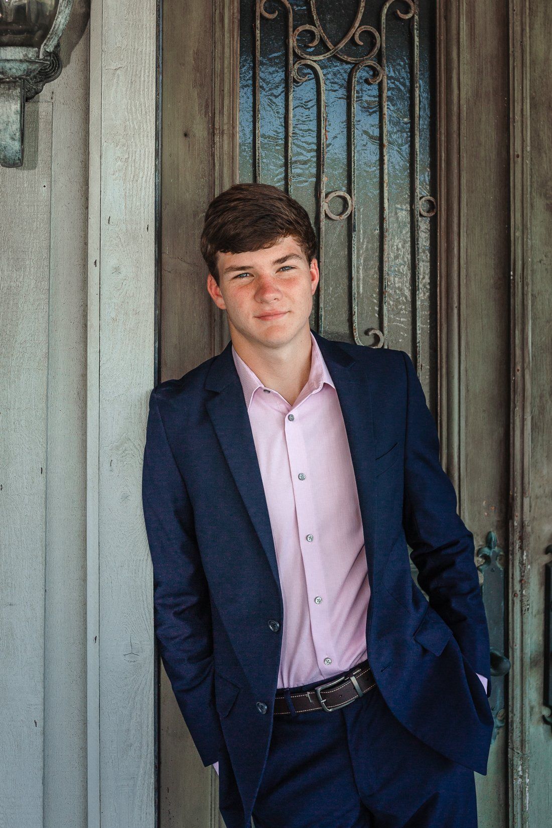 A young man in a suit and pink shirt is standing in front of a door.
