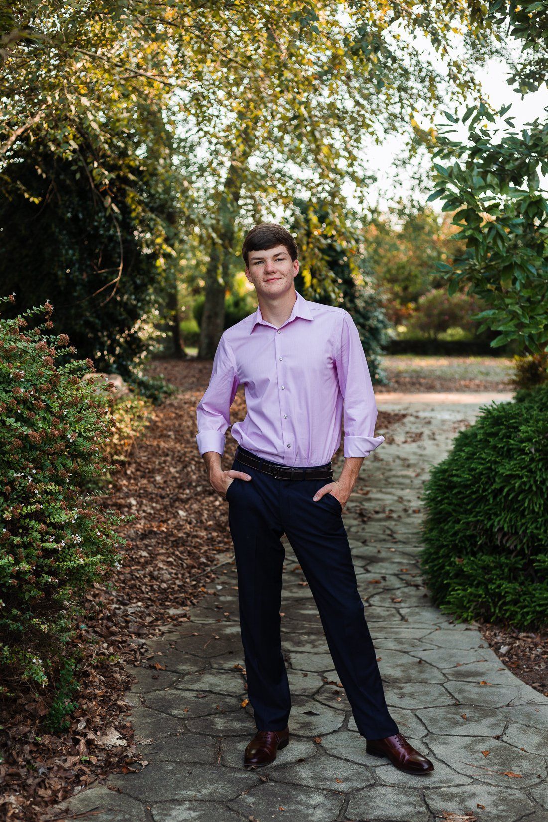 A young man in a purple shirt and black pants is standing on a stone path in a park.