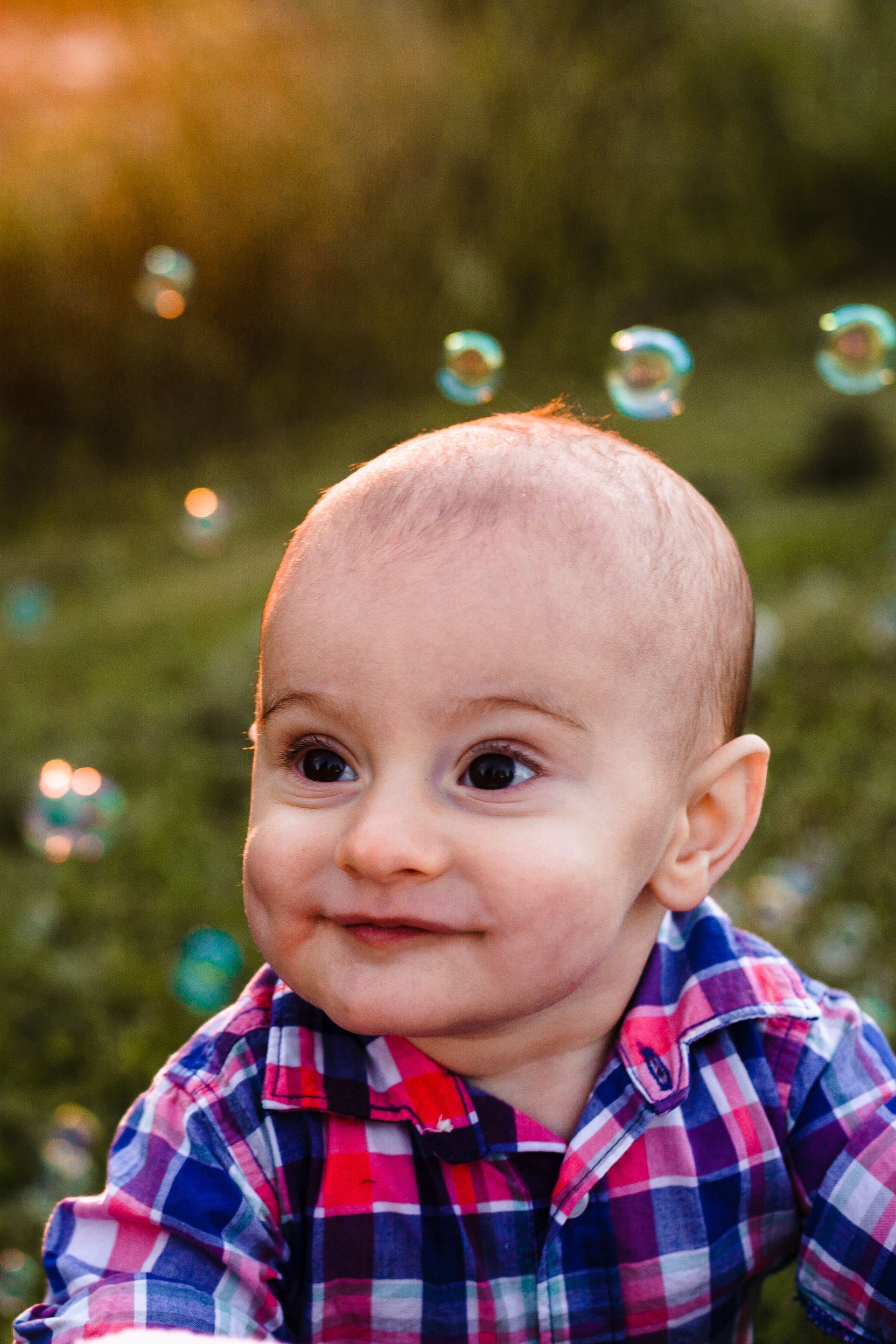 A baby in a plaid shirt is sitting in front of soap bubbles.