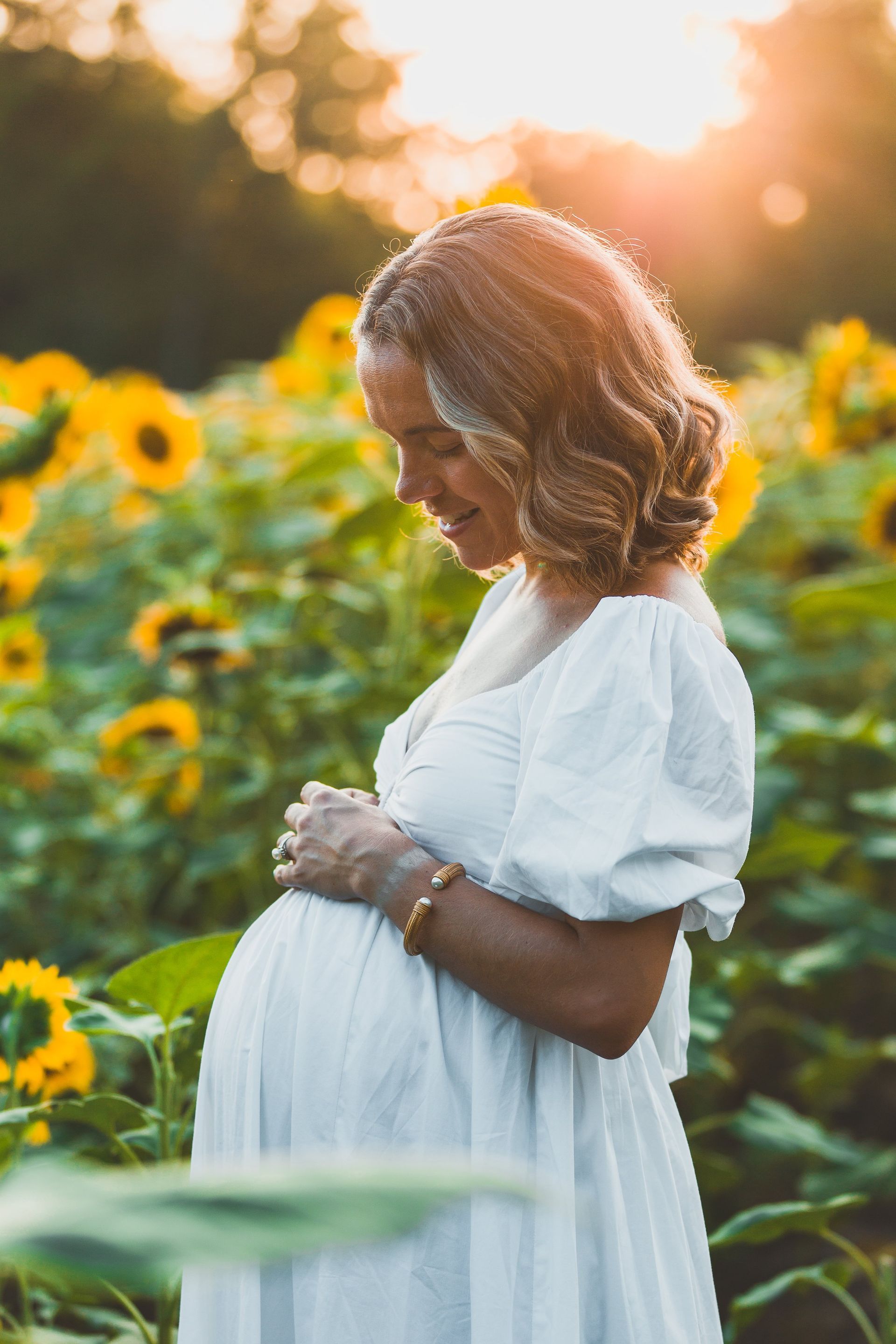 A pregnant woman in a white dress is standing in a field of sunflowers.