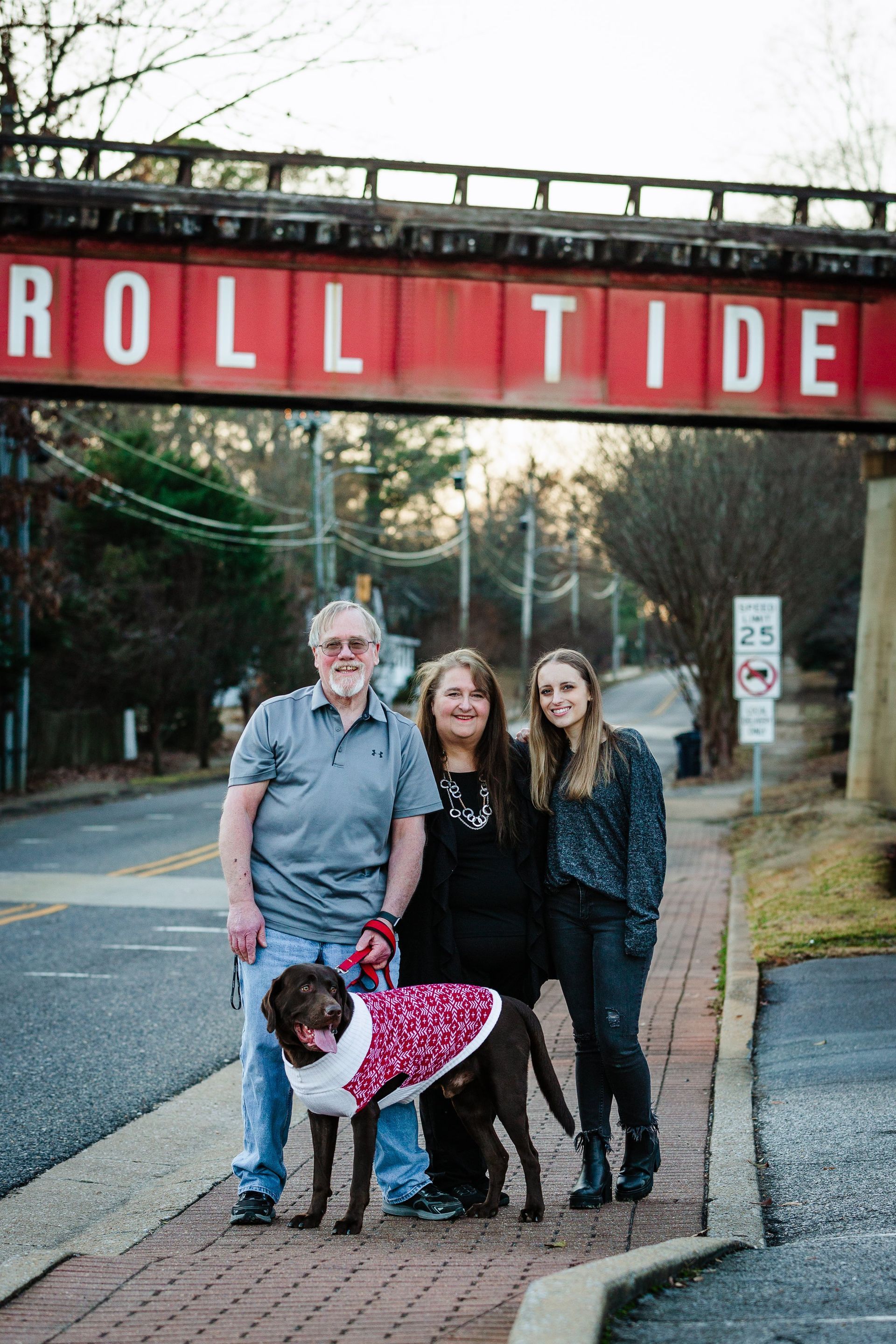A family is standing next to a dog on a sidewalk under a bridge.