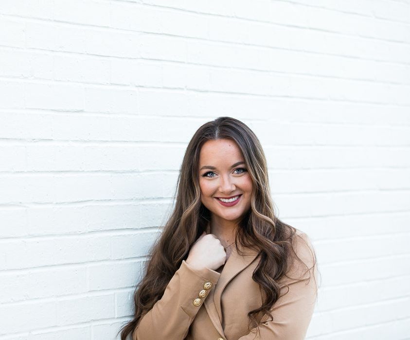 A woman in a tan coat is standing in front of a white brick wall.