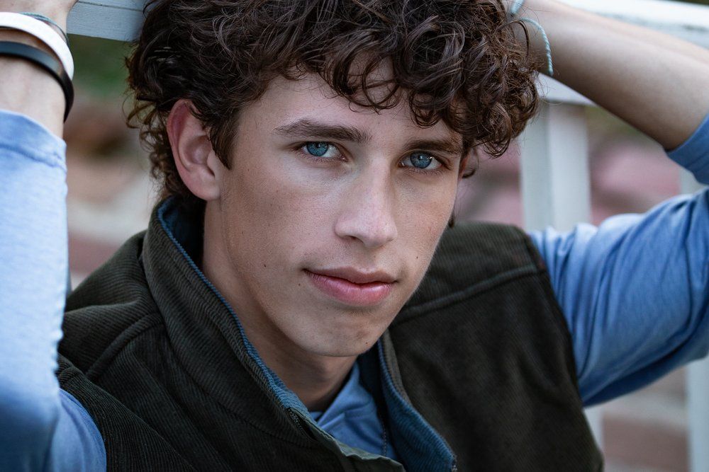 A young man with curly hair and blue eyes is leaning against a railing.