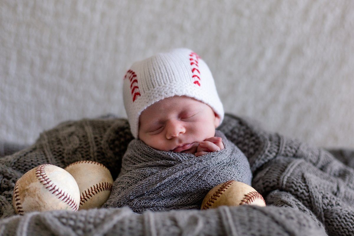A newborn baby wrapped in a blanket is sleeping in a basket with baseballs.