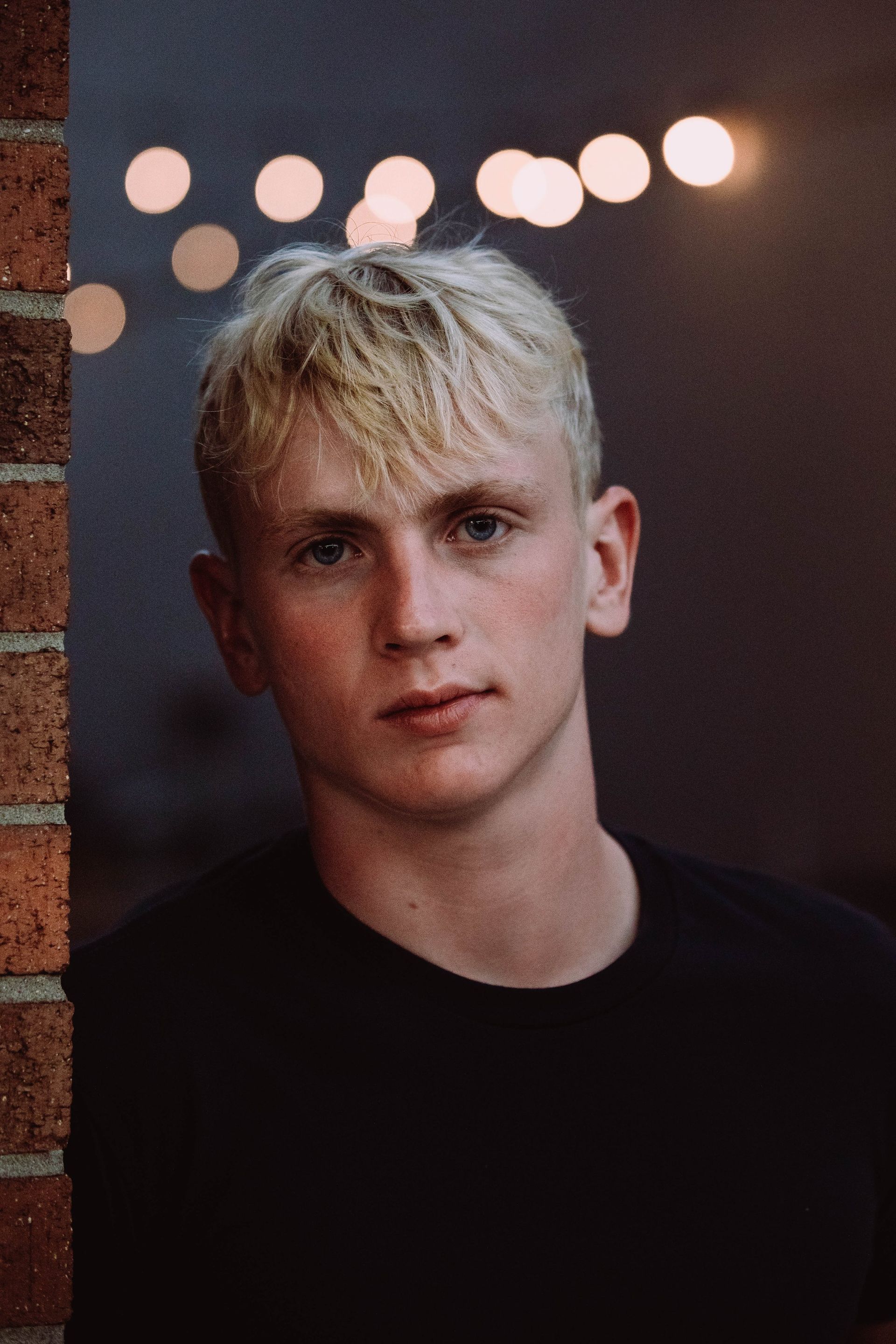 A young man with blonde hair is standing next to a brick wall.