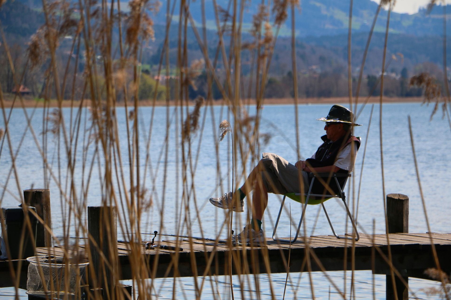 Person relaxing in a chair on a wooden dock by a lake with reeds and mountains in the background