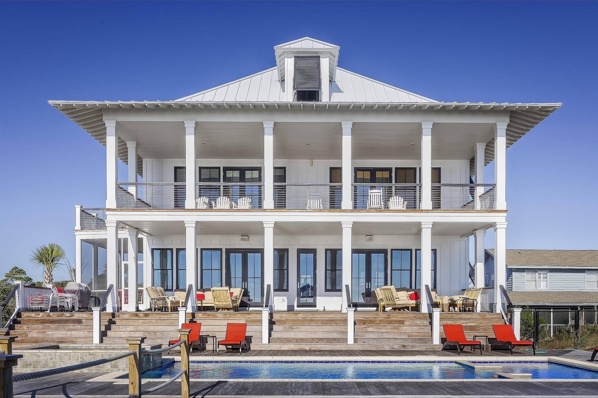 Large white waterfront house with wraparound porch and pool under a clear blue sky