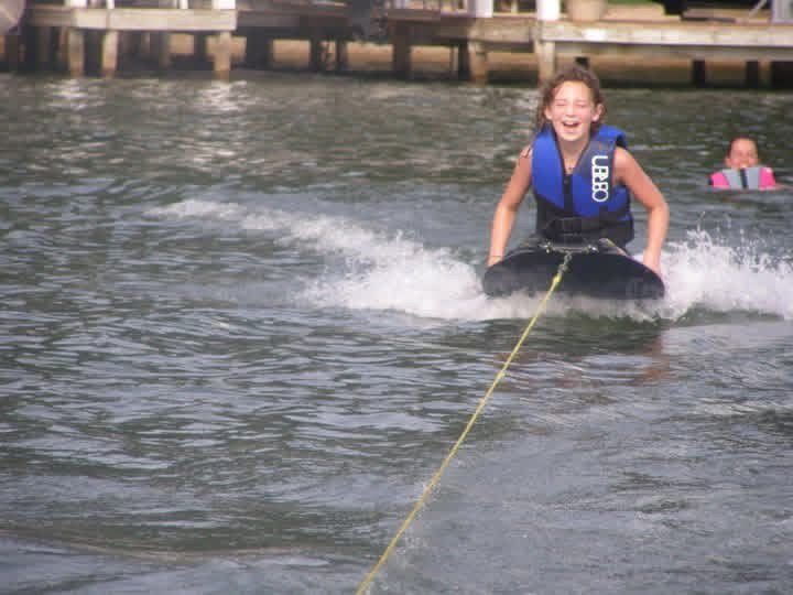 Person in a blue life jacket kneeboarding on water, being towed past a dock at Lake Gaston.