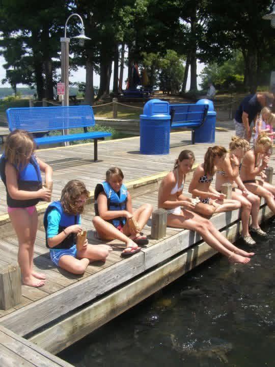 Children in life jackets sitting on a dock, dipping their feet into the water at Lake Gaston near a blue bench.