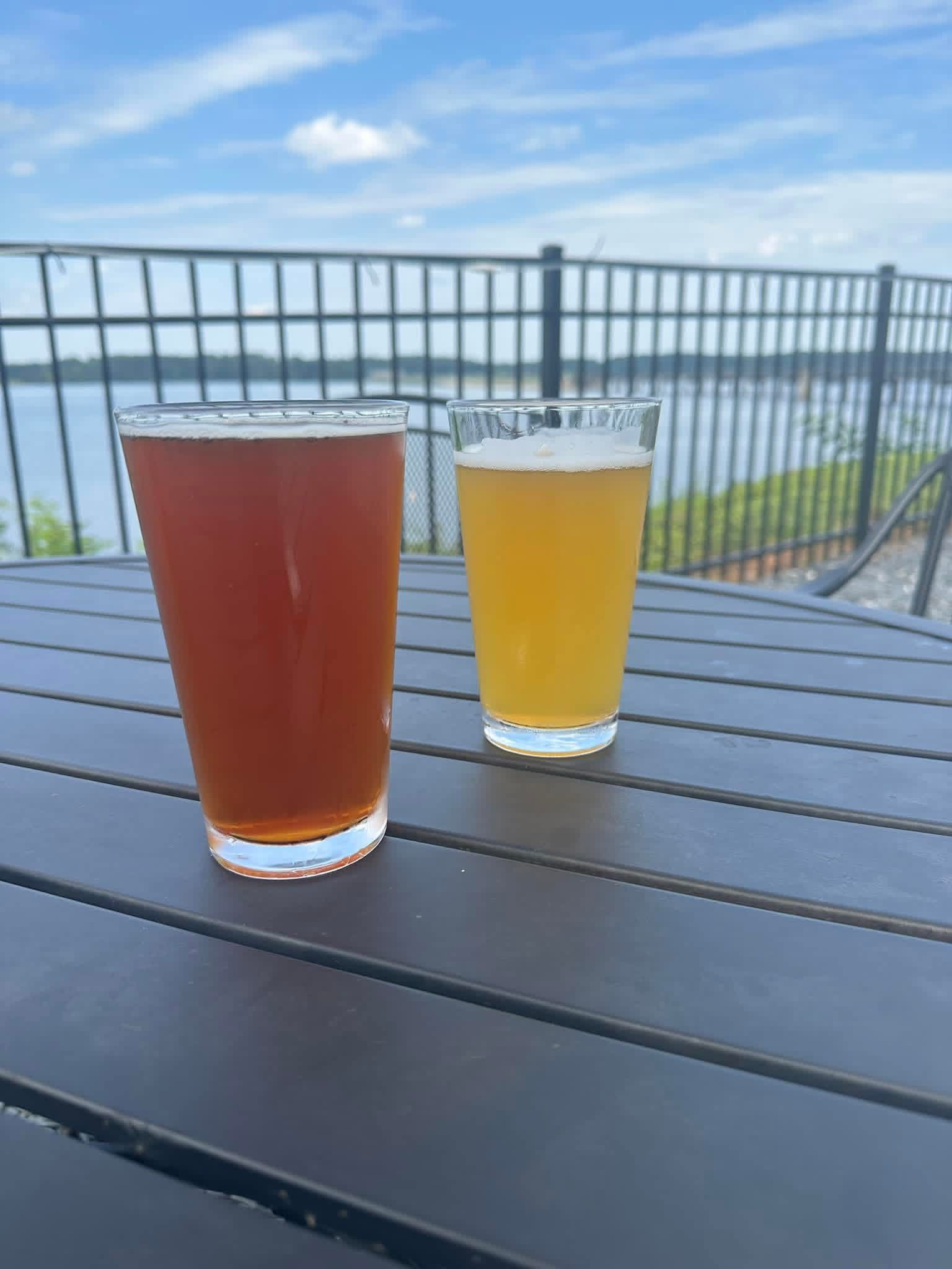 Two glasses of beer on a table by Lake Gaston waterfront railing under a blue sky,
