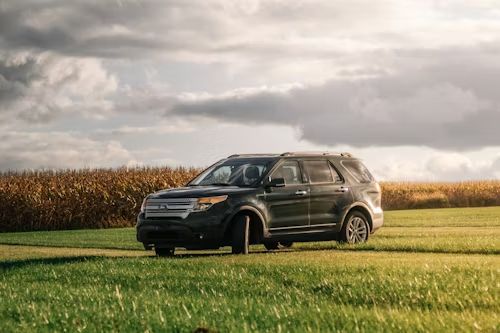 Black SUV driving through a grassy field with cornfield and cloudy sky in the background