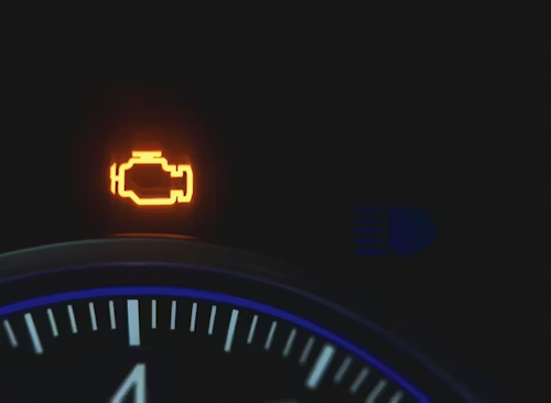An illuminated amber check engine light on a dark vehicle dashboard gauge cluster.