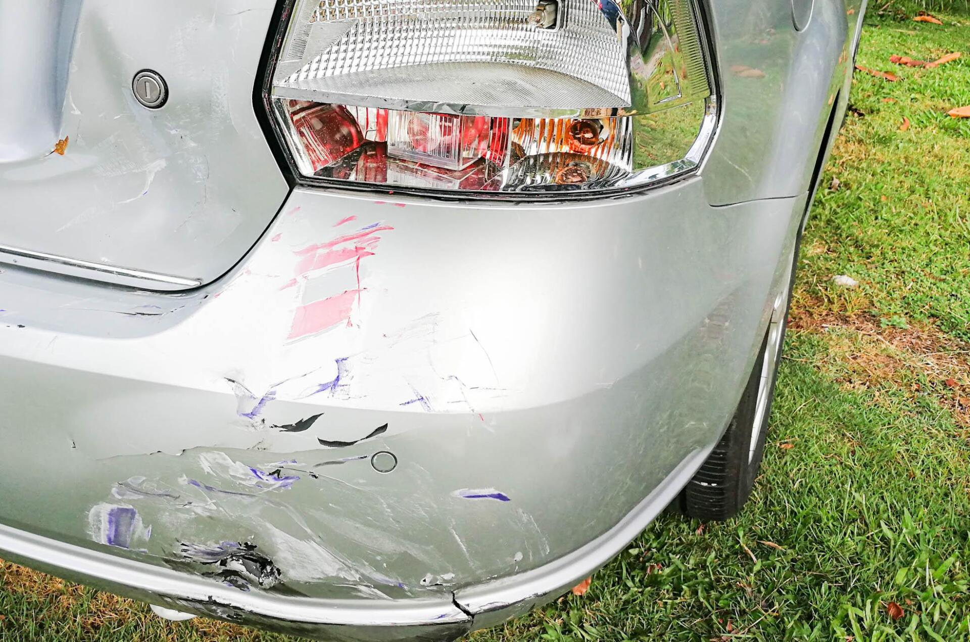 A silver car with a damaged bumper is parked in the grass.