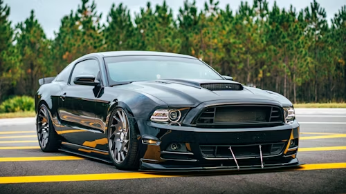 Black customized Ford Mustang sports car on a runway with a forest backdrop.