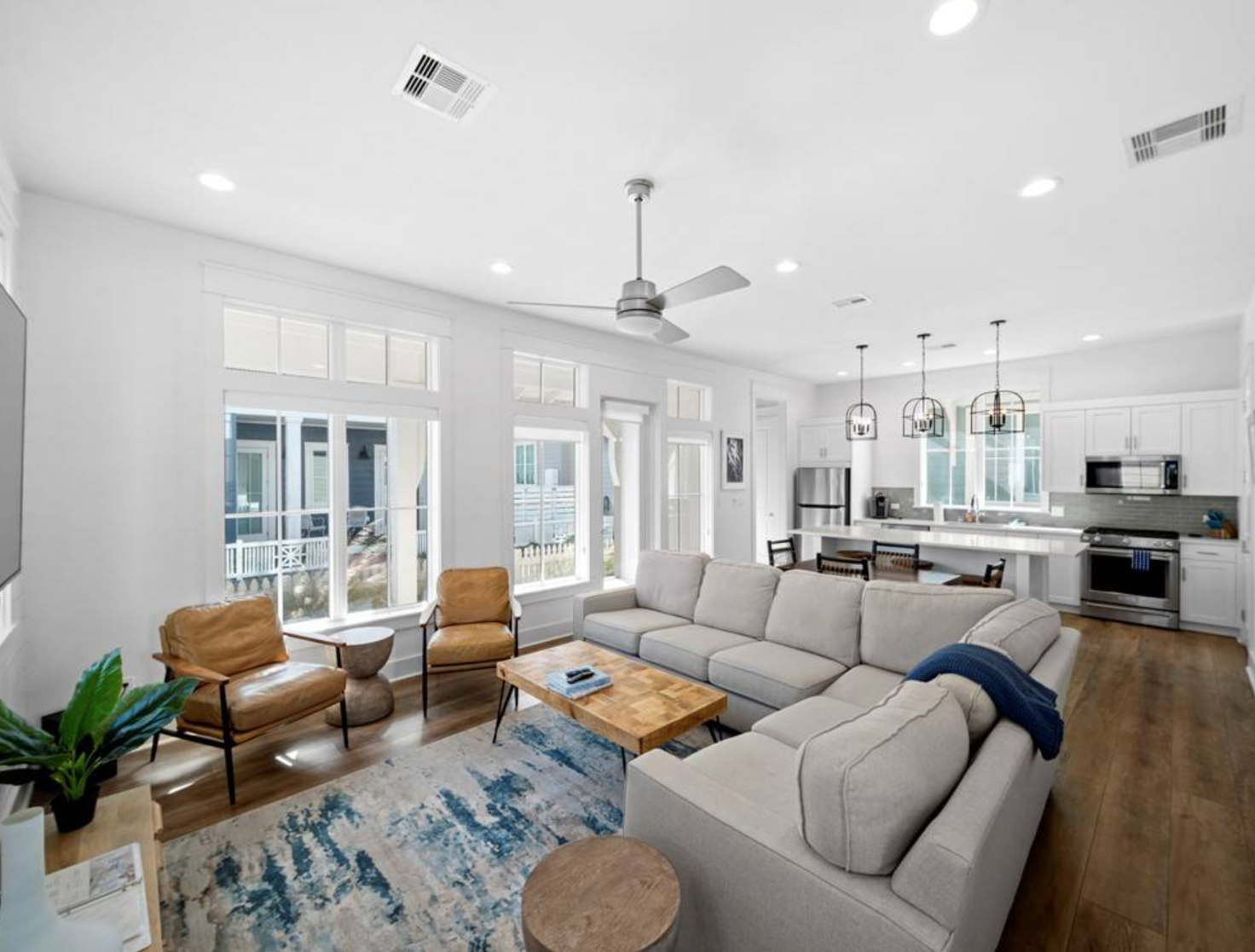 Spacious, bright living room with open kitchen. Light gray sectional, wooden coffee table, blue rug, and large windows.