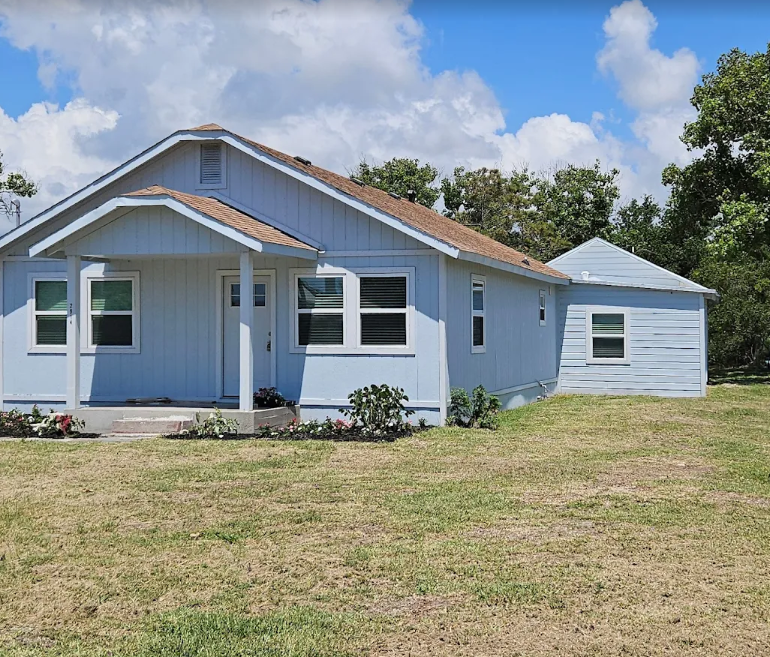 Light blue house with brown roof, white trim, and small front yard, under blue sky.