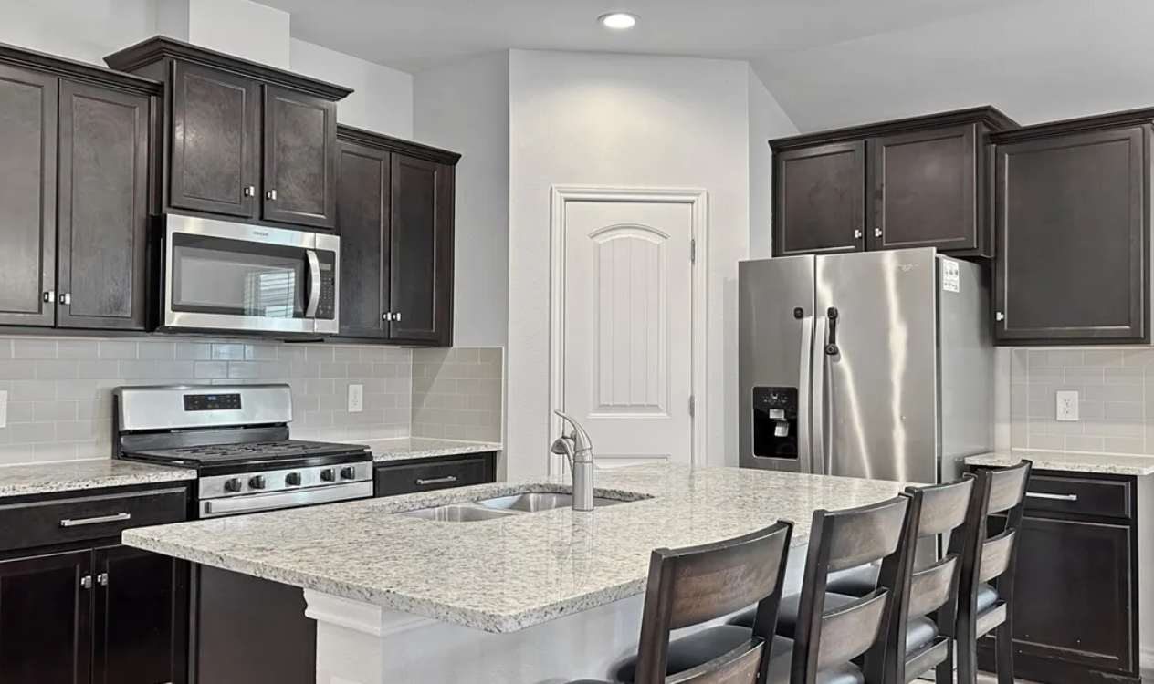 Kitchen with dark cabinets, granite island with stools, stainless steel appliances, and white walls.