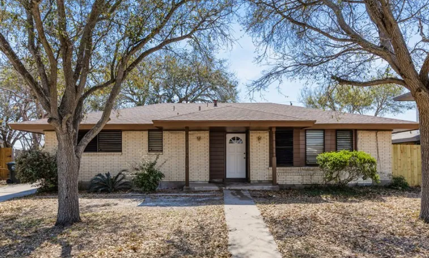 Single-story brick house with brown trim, centered walkway, trees in front yard, under a sunny sky.