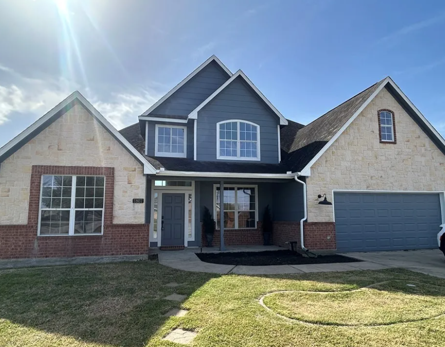 Two-story house with blue siding, brick, and tan stucco exterior under a sunny sky.
