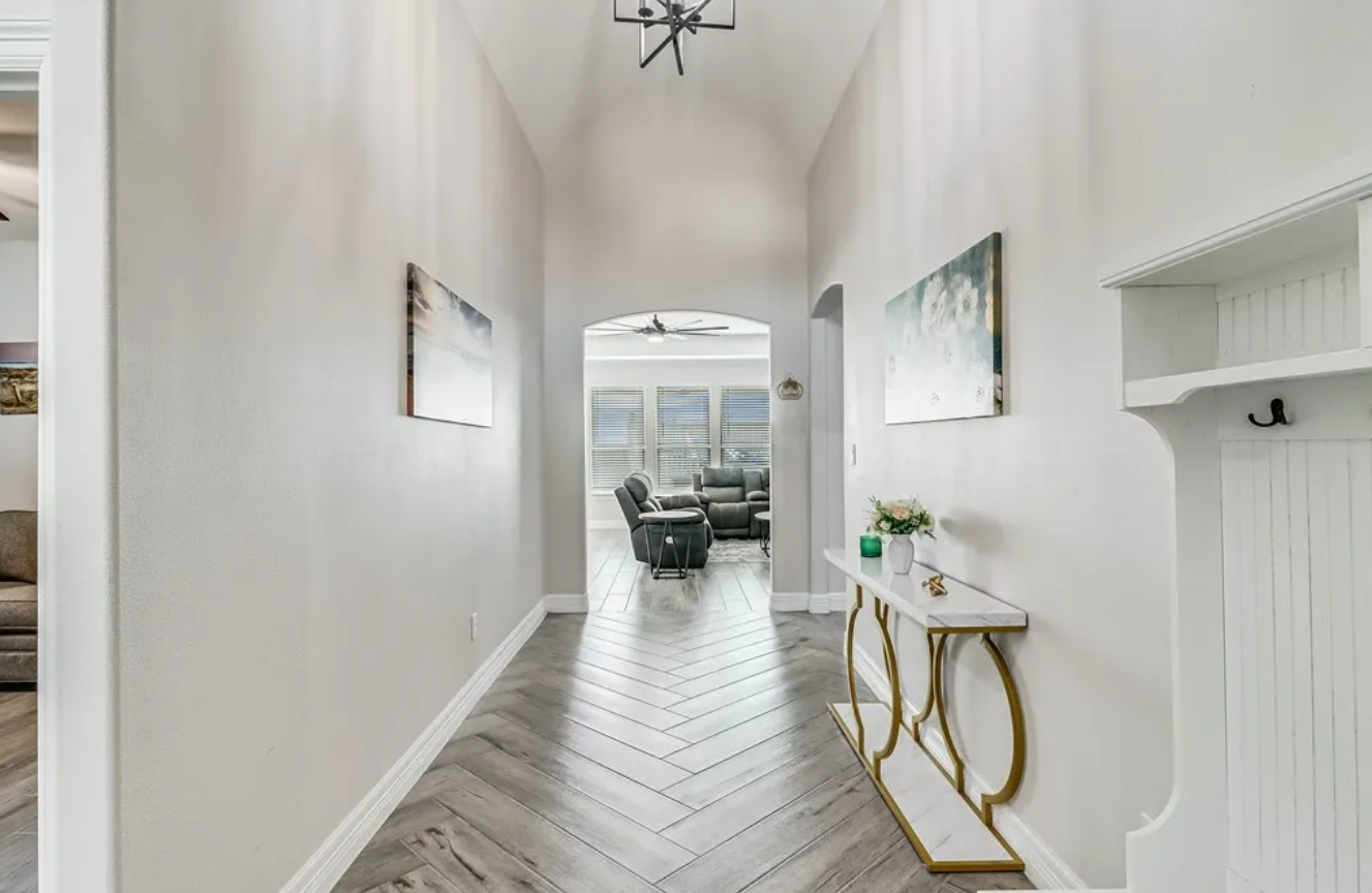 Hallway with light gray walls, herringbone floor, decorative table, and arched doorway leading to a living room.