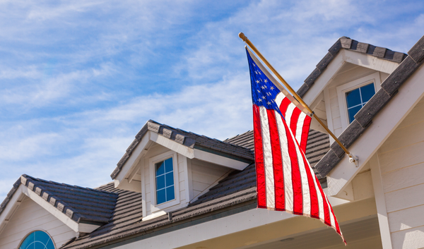 American flag waving outside a white house with dormers under a blue sky.
