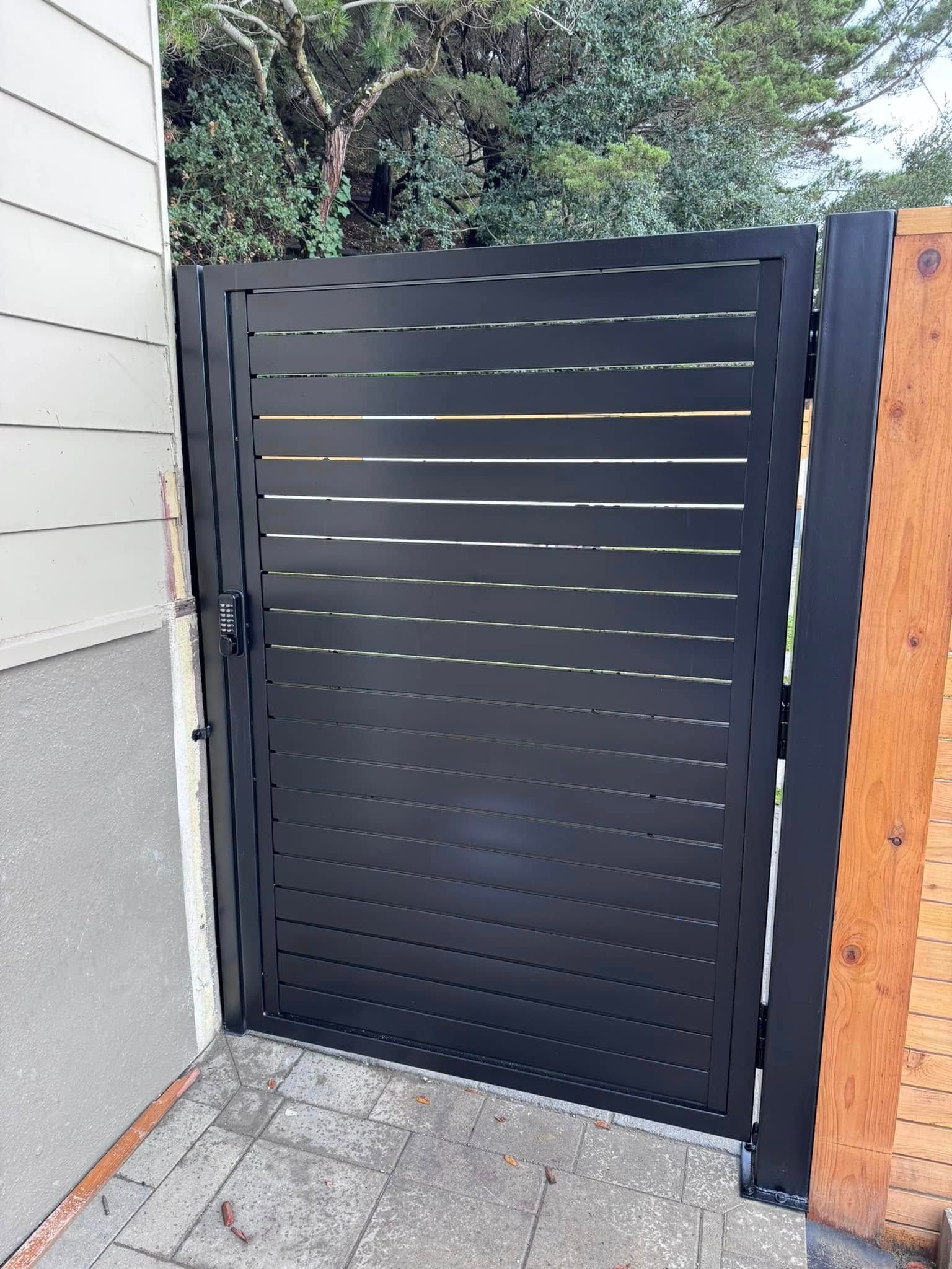 Black metal gate with horizontal slats, next to a house and wooden fence.