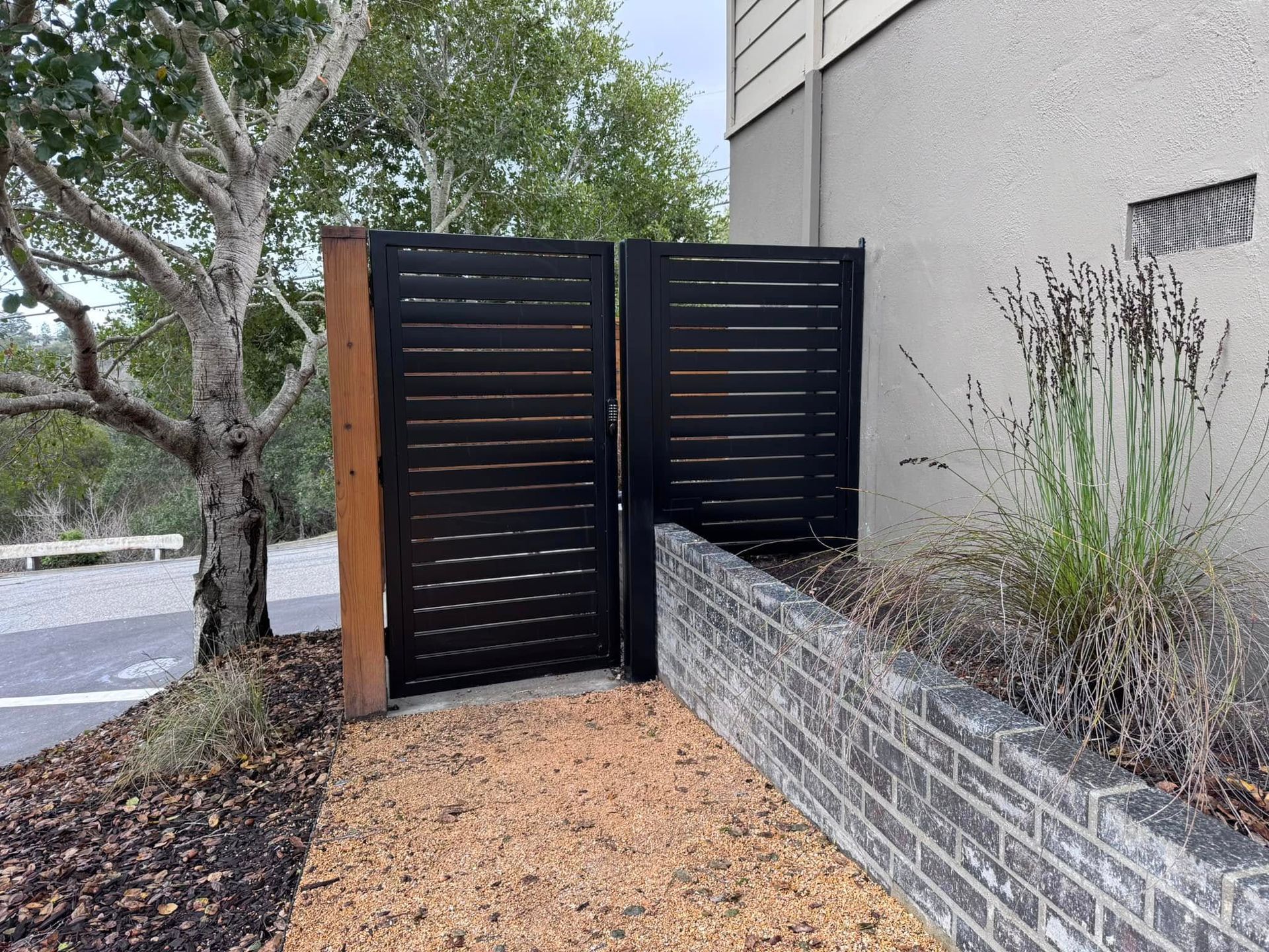 Black slat gate, brick wall, and gravel path near a building and tree.