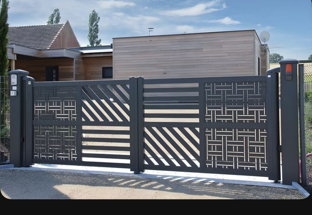 Dark gray metal driveway gate with geometric cut-out designs; in front of a light brown house.