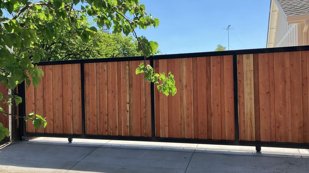 Wooden fence with black frame, on concrete, with tree branches and blue sky in the background.