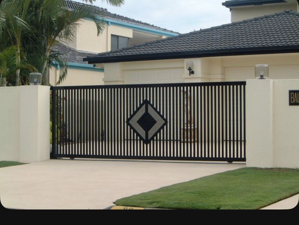 Black metal driveway gate with a diamond-shaped design set between cream-colored pillars, in front of a house.