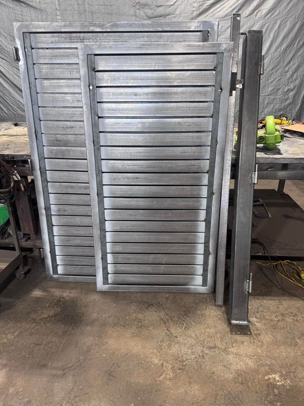 Metal gate components stacked on a concrete floor in a workshop. Gray metal frames with horizontal slats.