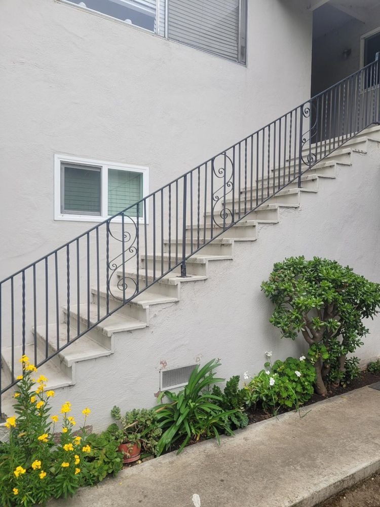 Exterior staircase with black railing, leading up to a building entrance, surrounded by plants and flowers.