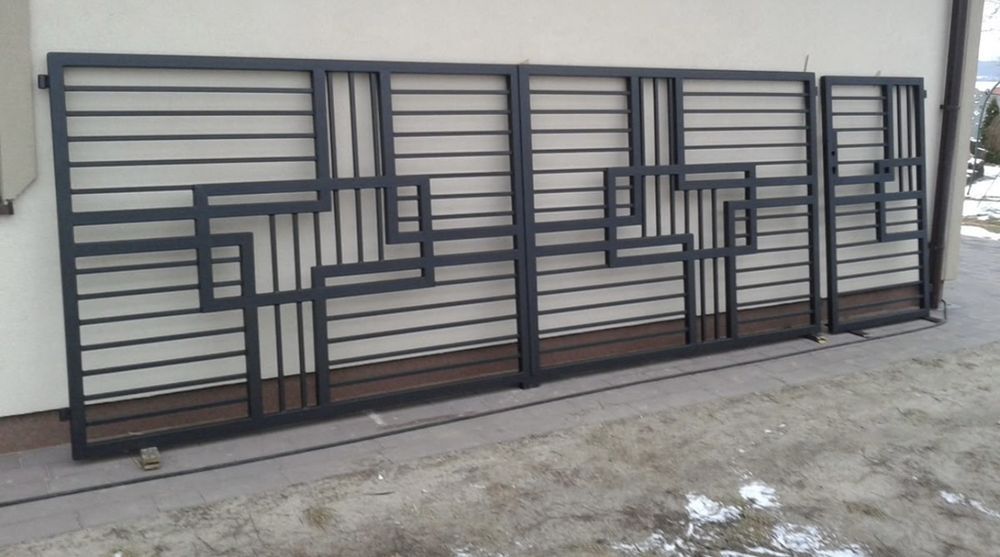 Black metal gates with geometric designs, set against a cream wall with horizontal siding, outdoors.