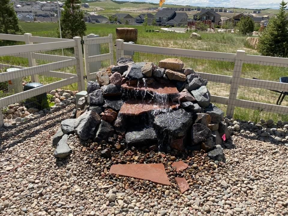 A small waterfall is surrounded by rocks and a white fence.