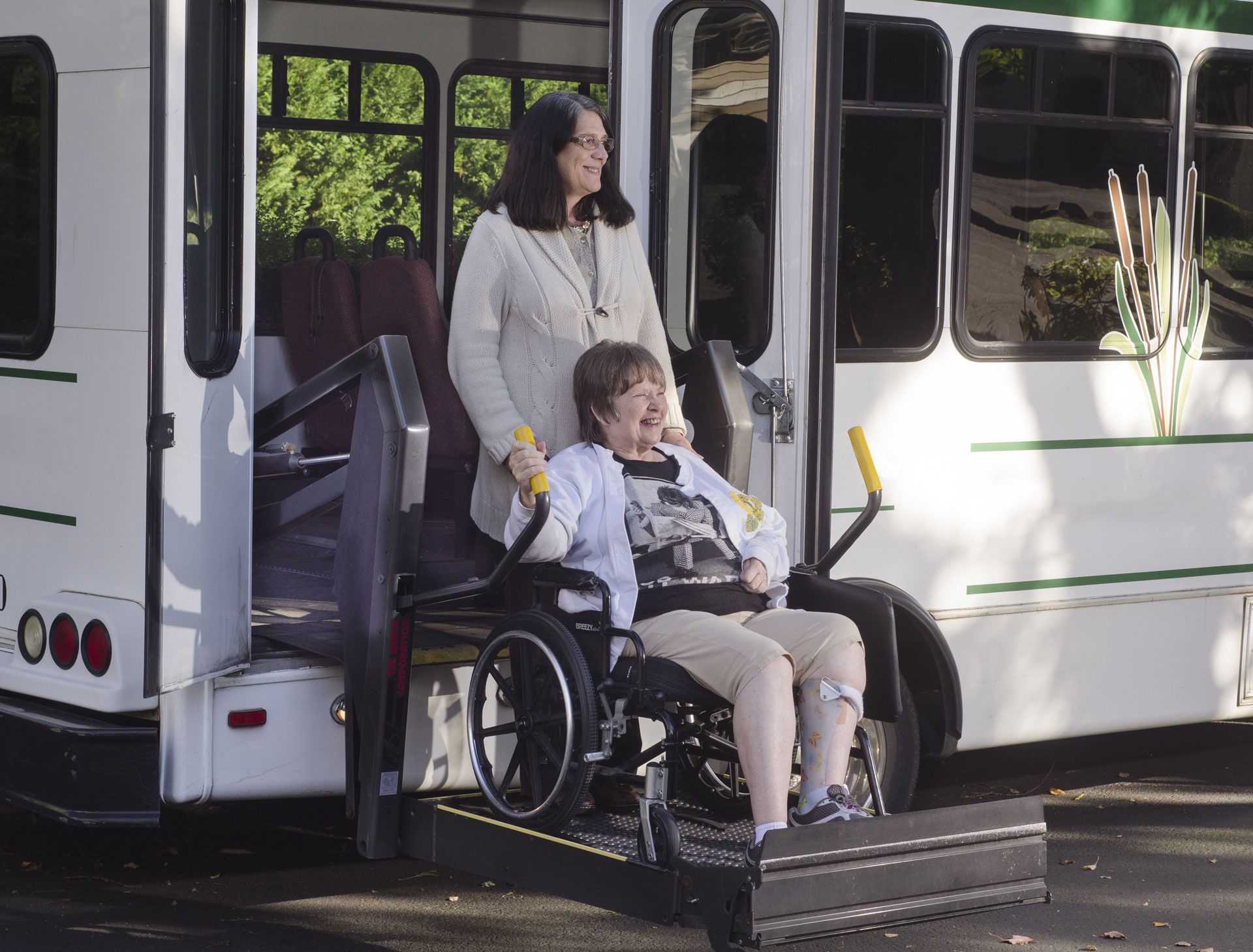 Woman assists person in wheelchair onto a bus with a lift. Outdoors, sunny.