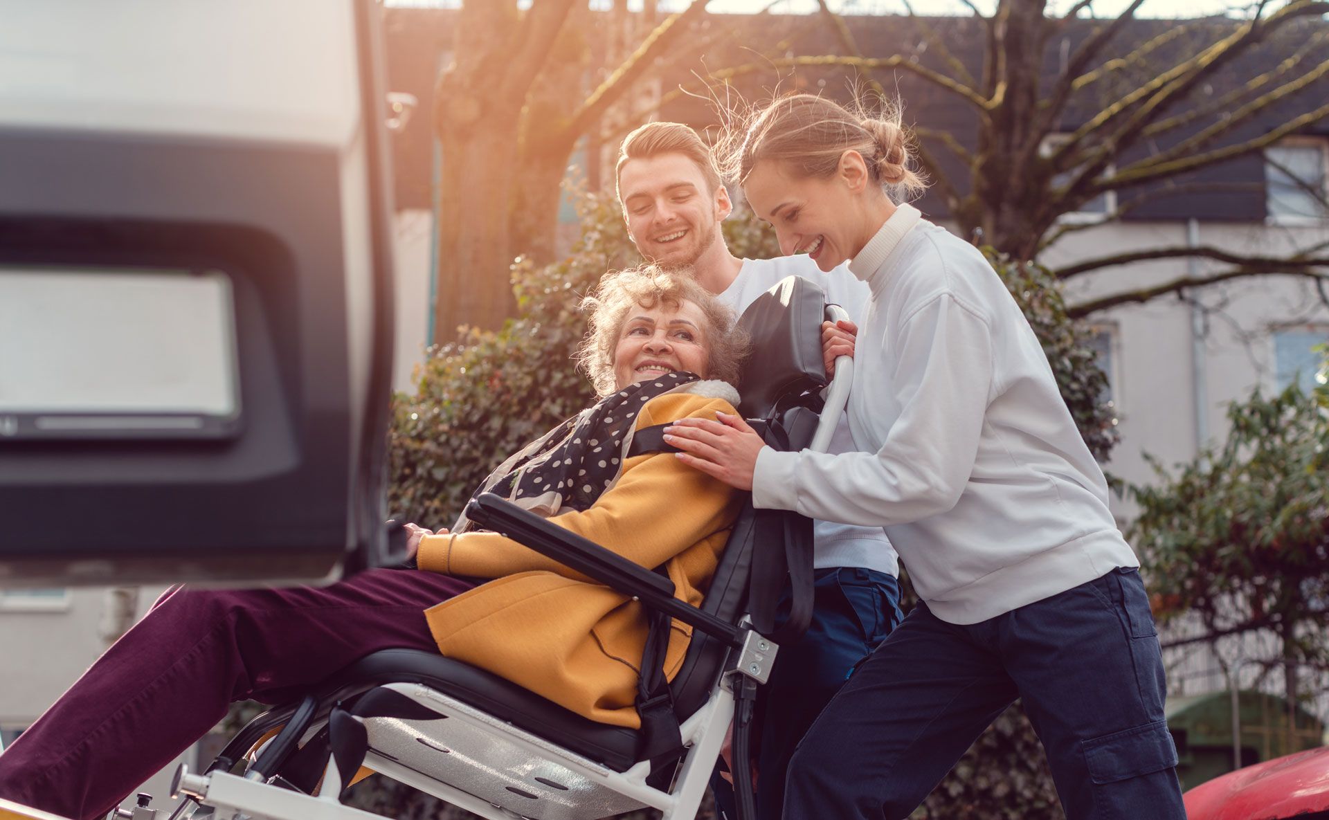 Family helps a woman in a wheelchair. Smiling, outdoors, sunlight, near a vehicle.