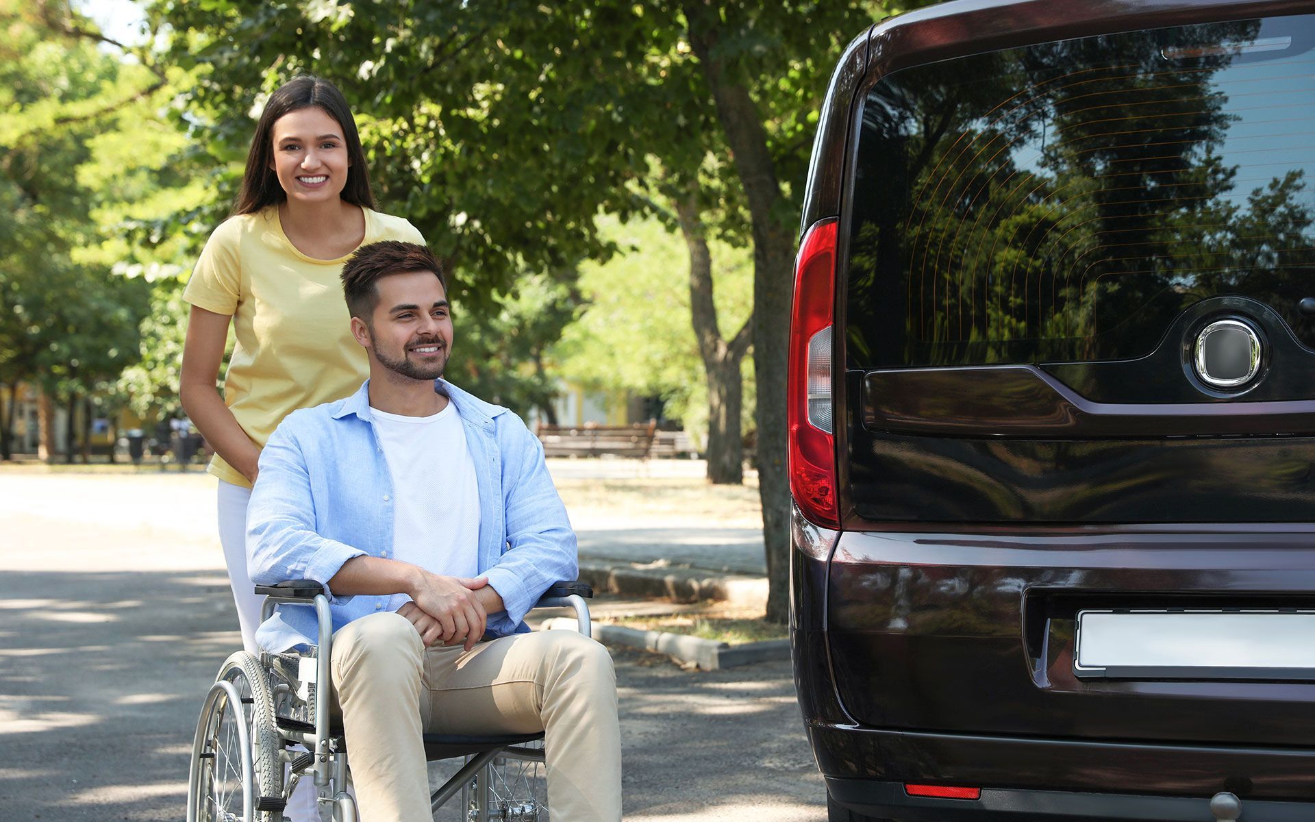 Woman pushing a man in a wheelchair towards a dark van in a park; both are smiling.