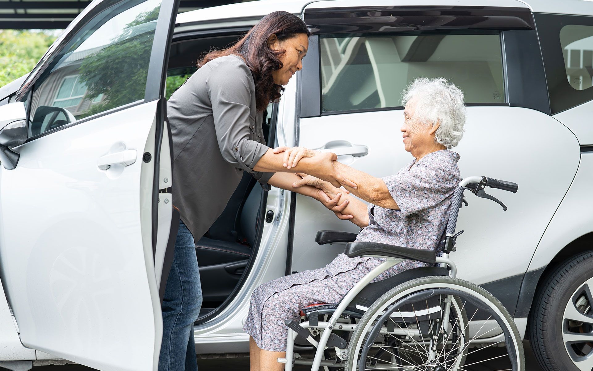 Woman helping elderly woman in wheelchair enter a silver car.