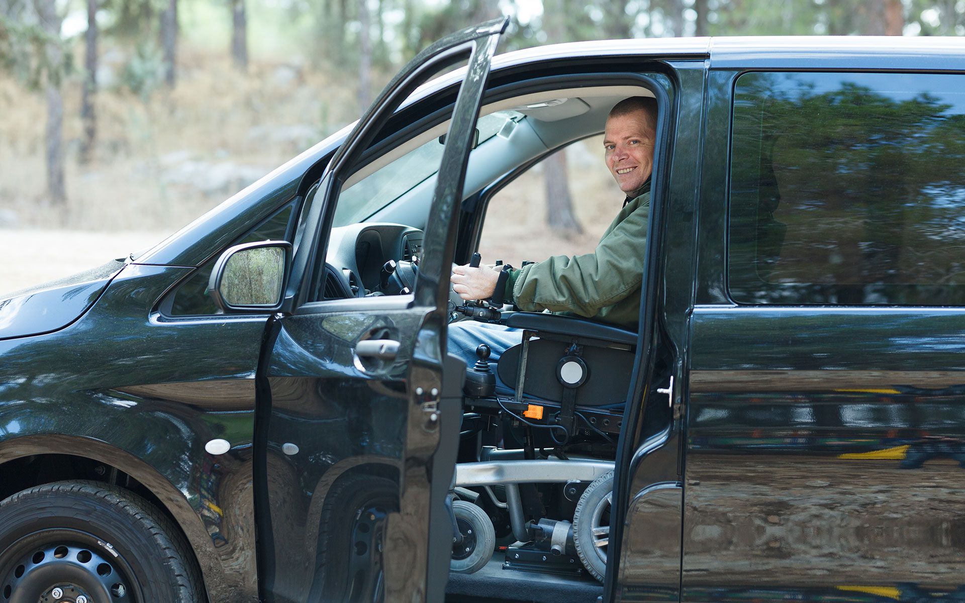 Man in a wheelchair drives a modified black van with the door open, smiling in an outdoor setting.