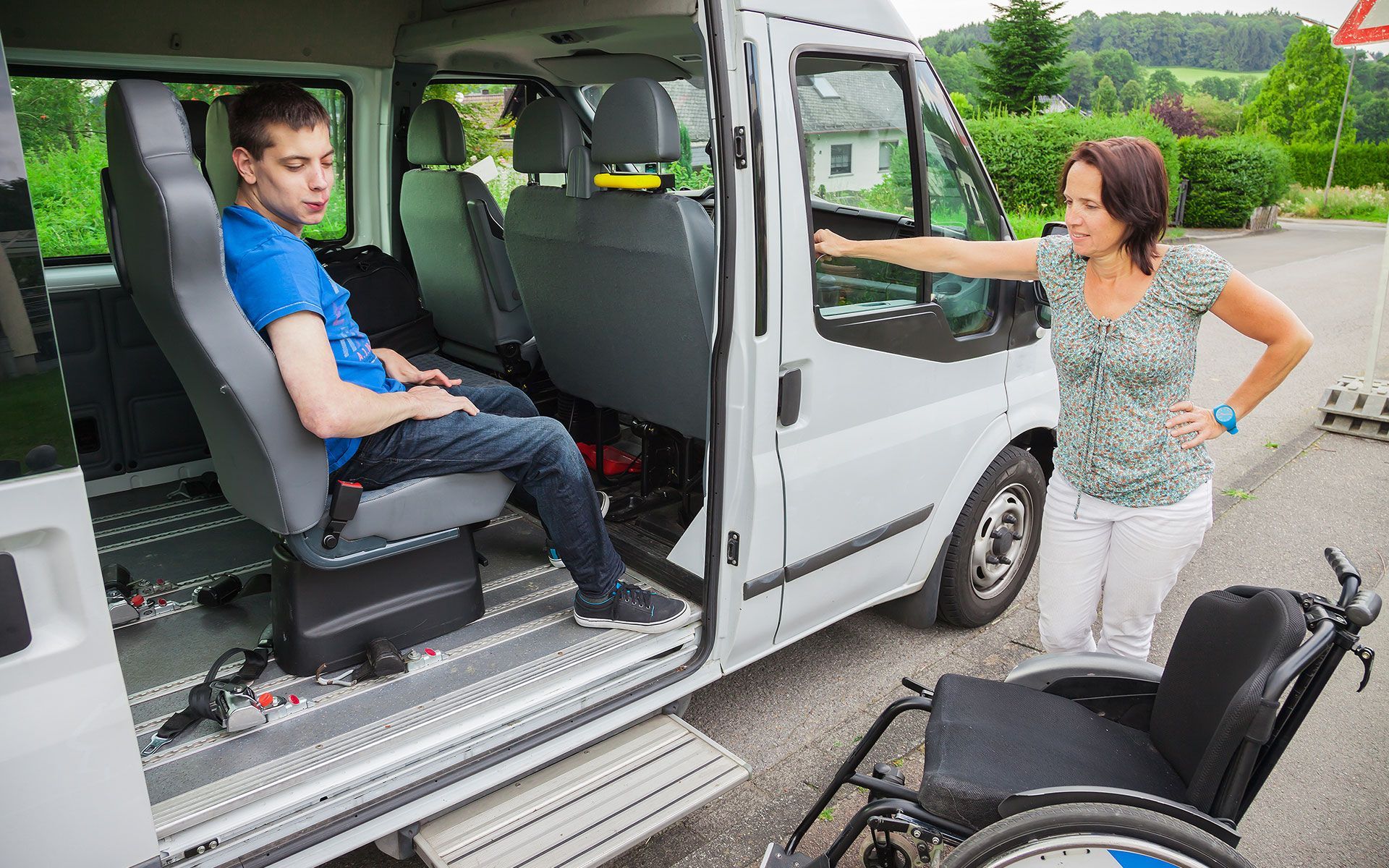A man in a van, seated, with a woman standing, wheelchair nearby. Outdoors, white van, daytime.