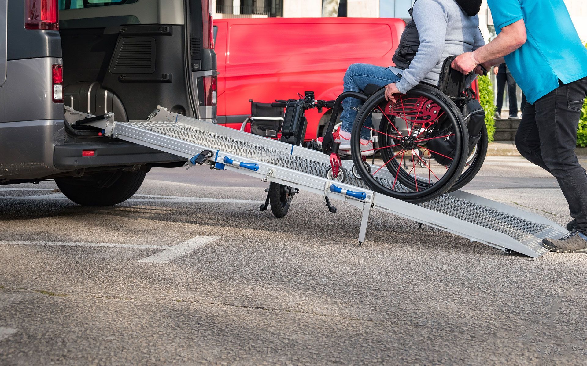 Person in wheelchair being assisted up a ramp into a red van by another person.
