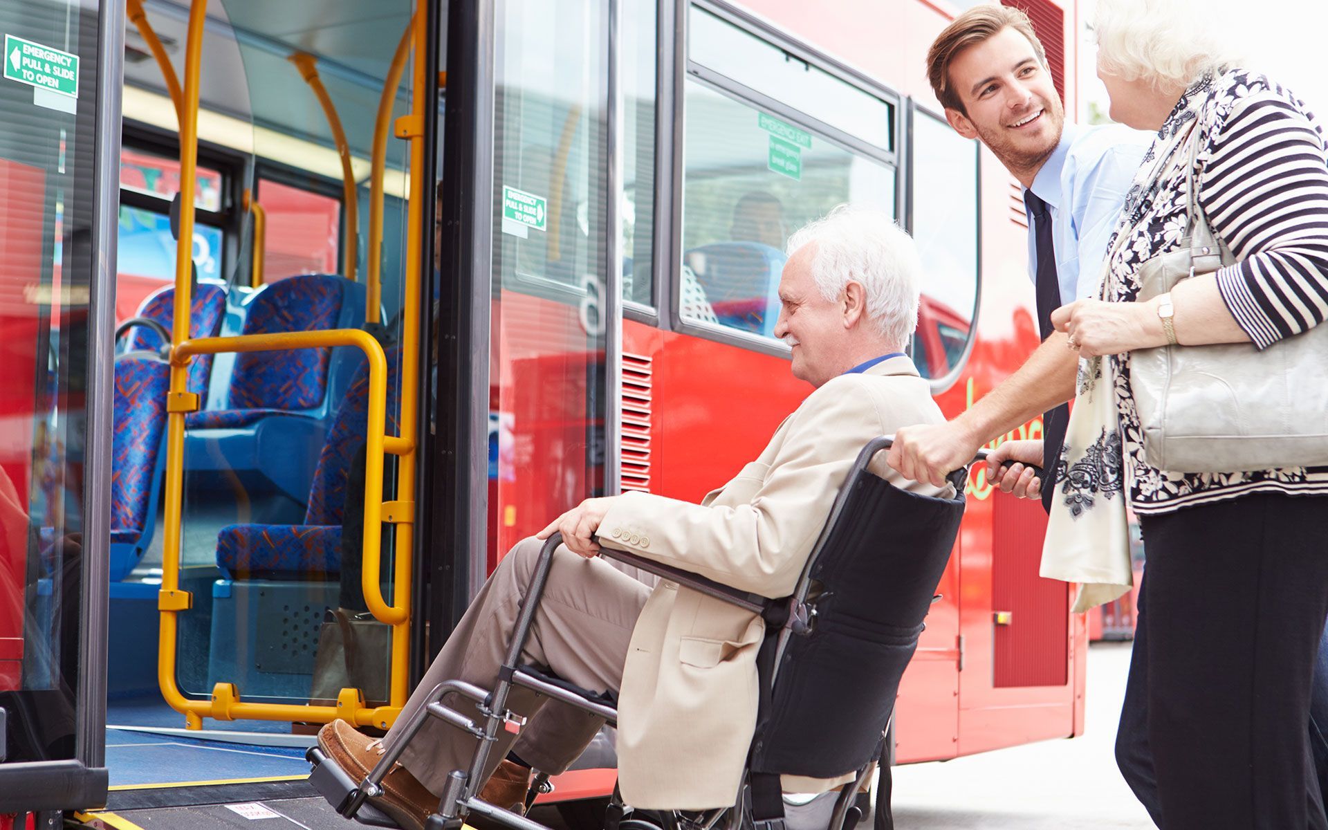 Man in wheelchair boarding a red bus with assistance from bus driver and woman.