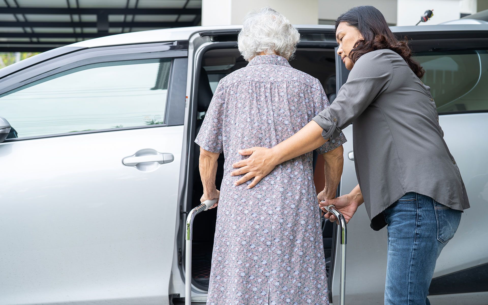Woman helps an elderly person with a walker get into a silver car.